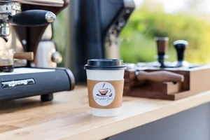 Disposable coffee cup on wooden counter near espresso machine and coffee tools.