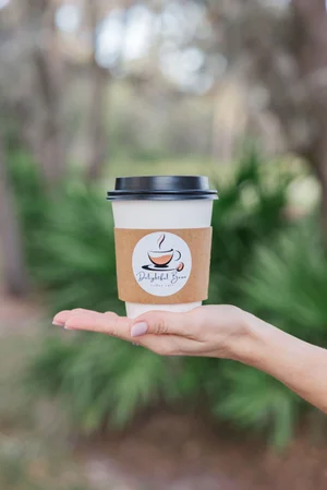 Hand holding a coffee cup with logo in a natural setting.