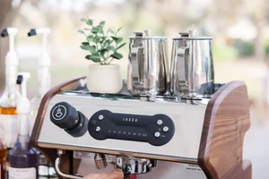 Espresso machine with stainless steel components and wooden side panels, topped with metal milk frothers and a small potted plant; bottles of syrup in the background.