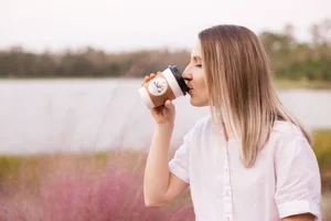 Woman drinking from a to-go coffee cup outdoors, wearing a white shirt, in front of a scenic lake background.