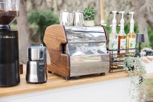 Espresso machine setup with a coffee grinder, milk frother, and syrup bottles on a wooden counter.
