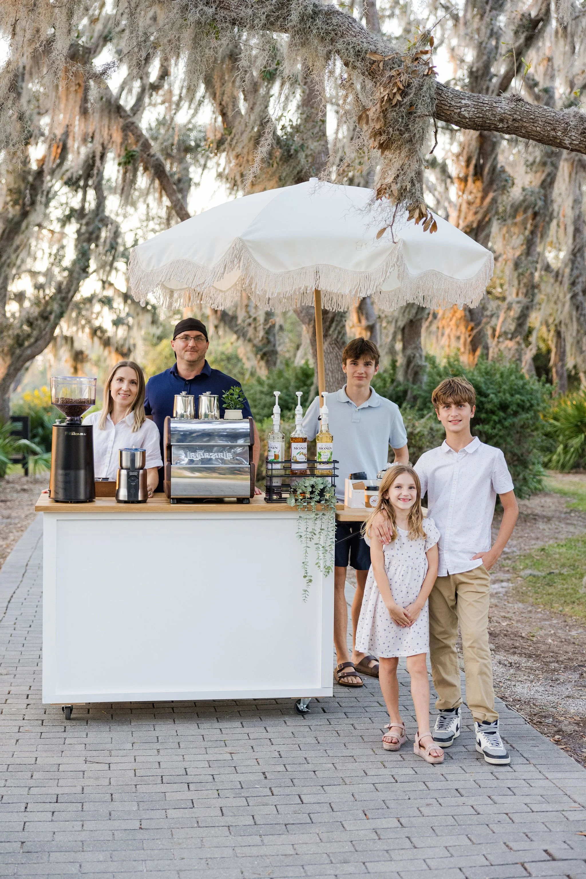Family standing behind outdoor mobile coffee cart with espresso machine and grinder under a fringed umbrella in a park.