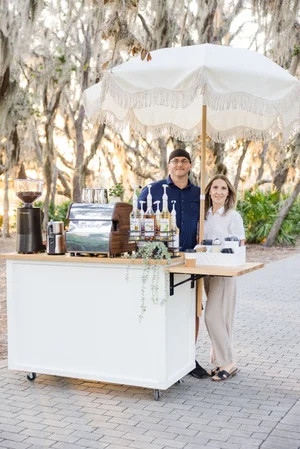 Mobile coffee cart with umbrella in outdoor setting, man and woman standing behind, coffee machine and syrups on cart.