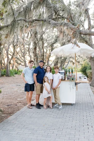 Family posing outdoors in wooded area, near a mobile coffee cart with drinks and umbrella, on a paved path under draped tree branches.