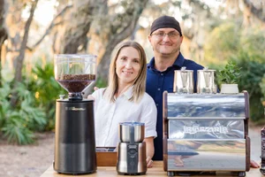 Two people standing behind coffee equipment outdoors with trees in the background.