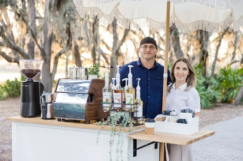 Outdoor coffee stand with a man and woman posing behind it, featuring a La Marzocco espresso machine, coffee grinder, Monin syrup bottles, and disposable coffee cups, set in a park with trees in the background.