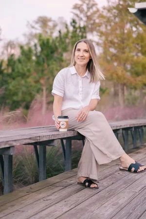 Woman sitting on a wooden bench outdoors, holding a coffee cup, wearing a white shirt and beige pants.